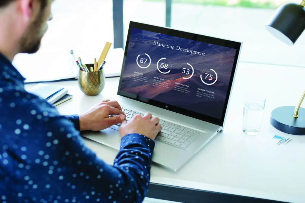 A man working on a silver HP laptop displaying 'Marketing Development' charts in a bright, modern home office setting